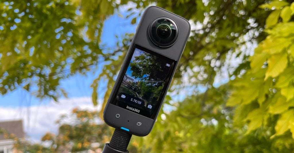 Detailní záběr na 360° kameru Insta360 umístěnou na černé tyči. Na displeji kamery je vidět živý náhled na stromy a budovy s údajem o natáčení v rozlišení 5.7K/30. Pozadí tvoří silně rozostřené zelené listy stromů proti modré obloze.