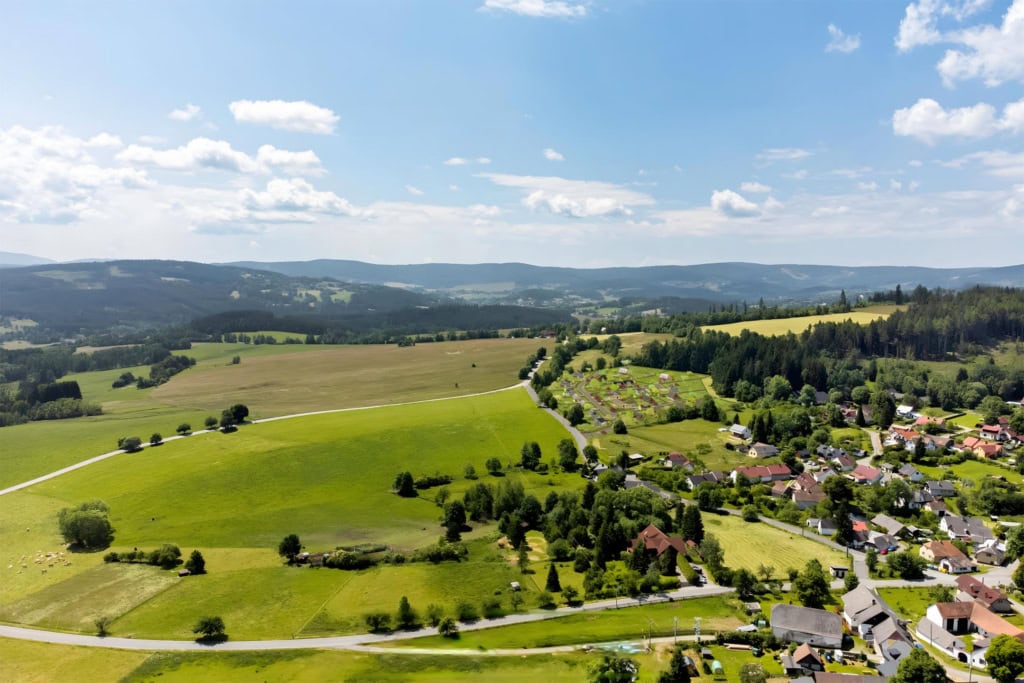 Široký letecký pohled na šumavskou krajinu a stávající vesnici. Do zelených luk na okraji obce je plynule zasazena vizualizace nové zástavby rodinných domů. V pozadí jsou zalesněné kopce.